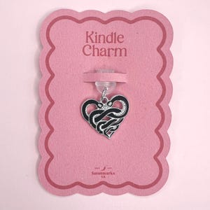 Può includere: Un ciondolo a forma di cuore con un design a serpente bianco e nero, appeso a una carta rosa. La carta presenta il testo "Kindle Charm" e il marchio "Solutmarks UK". Il ciondolo è attaccato a un supporto di plastica trasparente.