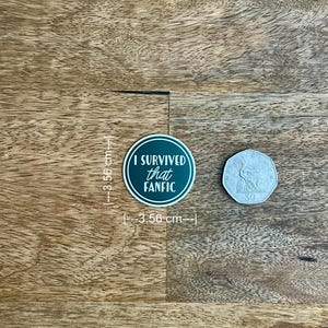 May include: A round sticker, 3.56 cm in diameter, with the text "I SURVIVED that FANFIC" in white on a dark green background. A 50-cent coin is next to the sticker. The sticker is on a wooden surface.