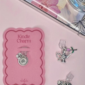 May include: A pink card with the text "Kindle Charm" displays a silver charm with a heart and a round charm. Two additional charms are visible: a teapot with a pink rose and a teacup. The background is a light pink surface.