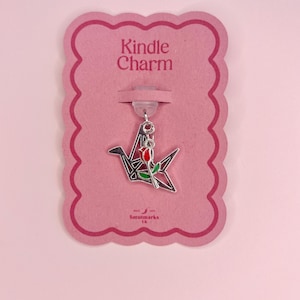 Puede incluir: Un charm plateado con un diseño de grulla de papel con una rosa roja y hojas verdes. El charm está sujeto a una tarjeta rosa con las palabras "Kindle Charm" y el logotipo de Smutmarks UK.