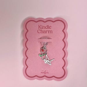 Puede incluir: Un charm plateado con una rosa roja y verde y una grulla de papel, sujeto a una tarjeta rosa con el texto "Kindle Charm". La tarjeta tiene un borde festoneado y el nombre de la marca "Smotmarks" en la parte inferior.