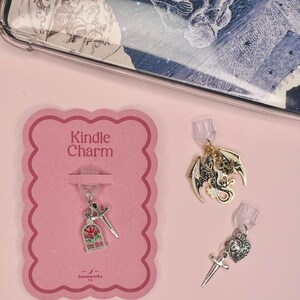 Op de afbeelding: Een roze kaart met de tekst "Kindle Charm" en drie bedels. Eén bedel heeft een roos en een zwaard, een andere een draak en de derde een hart en een zwaard. De bedels zijn zilver en goud.