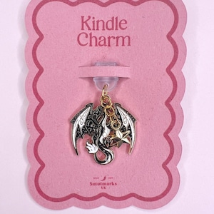 May include: A Kindle charm with a dragon design, featuring black and silver wings, a white body, and a gold star charm. The charm hangs from a clear plastic piece attached to a pink card with the words "Kindle Charm".