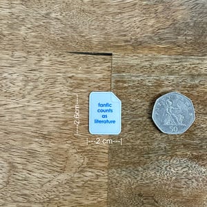 May include: A light blue sticker, 2.6 cm tall and 2 cm wide, with the text "fanfic counts as literature". A 50-cent coin is next to the sticker. The sticker and coin are on a wooden surface.