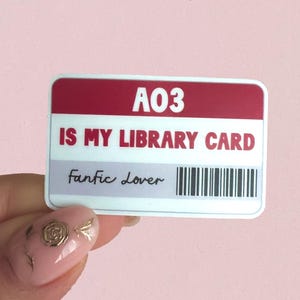 May include: A rectangular sticker with a red top section and white text that reads "A03 IS MY LIBRARY CARD." Below, the text "Fantic Lover" is written in cursive, with a barcode on the right. The sticker is held against a pink background.
