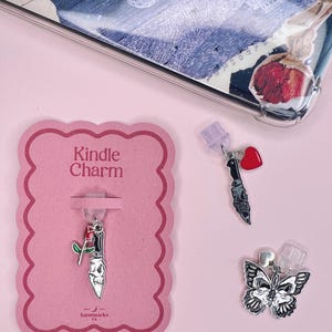 Puede incluir: Una tarjeta rosa con el texto "Kindle Charm" muestra un dije plateado con un cuchillo de calavera y una rosa roja. Otros dos dijes incluyen un cuchillo con una calavera y un corazón rojo, y una mariposa plateada. El fondo es rosa claro.