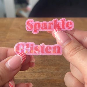 May include: Two pink, rectangular stickers with the words "Sparkle" and "Glisten" in a bubble font. The stickers have a glittery effect and are held up by a person's hands.