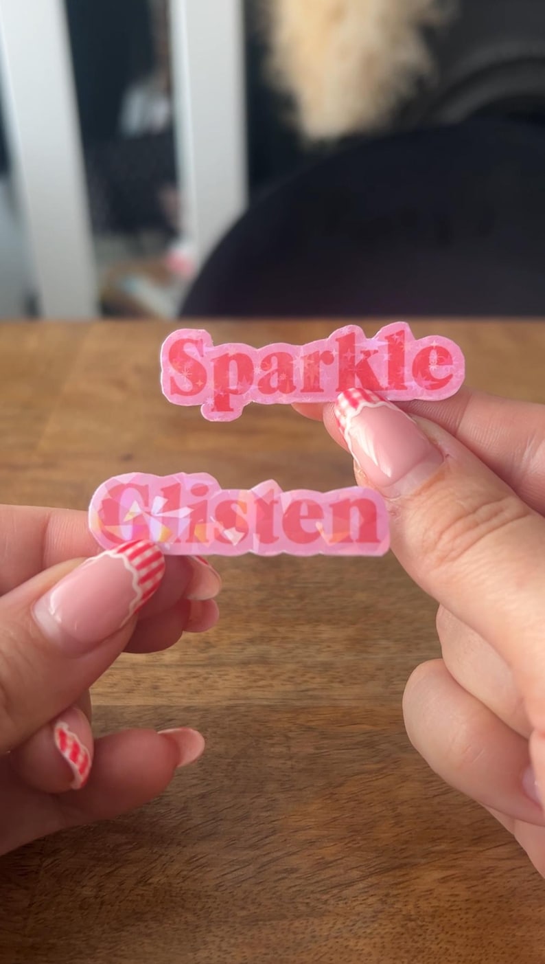 May include: Two pink, rectangular stickers with the words "Sparkle" and "Glisten" in a bubble font. The stickers have a glittery effect and are held up by a person's hands.
