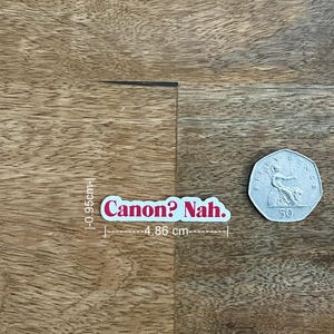 May include: A white sticker with the text "Canon? Nah." in pink lettering. The sticker is 0.37 inches tall and 1.91 inches wide. A 50-cent coin is next to the sticker on a wooden surface.