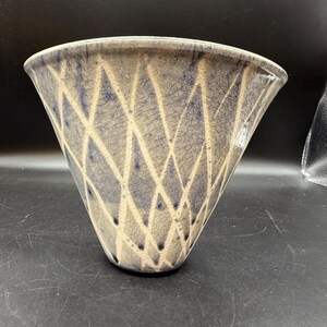 May include: A ceramic vase with a wide, flared opening and a conical shape. The vase features a pattern of intersecting lines in a cream colour against a blue-grey background. The vase has a textured surface and a slightly irregular rim.