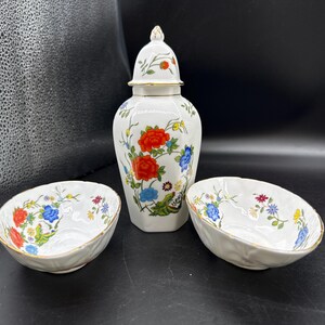 Pode incluir: Um conjunto decorativo de porcelana branca com um jarro com tampa e duas pequenas tigelas. As peças são adornadas com um padrão floral em vermelho, azul e amarelo. O jarro tem uma borda dourada e um design floral correspondente na tampa.