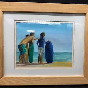 以下が含まれることがあります： ビーチでサーフィンをする人々の額入り絵画。3人の人物が水辺に立ち、サーフボードを持っています。絵画は青い海と空、砂浜を描いています。フレームは明るい色の木材で作られています。