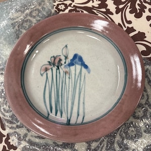 May include: A round ceramic plate with a mauve rim and a white center. The plate features a hand-painted design of blue and pink flowers with green stems. A thin blue line separates the rim from the center.
