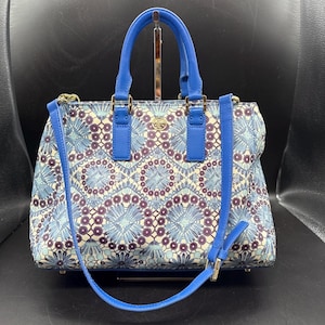 Puede incluir: Bolso estampado en azul y blanco con un diseño floral. Tiene un asa azul y una correa de hombro azul desmontable. Los herrajes dorados y un logotipo dorado son visibles en la parte delantera del bolso.