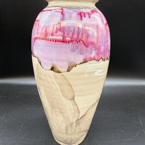 Könnte beinhalten: Eine hohe, handgefertigte Keramikvase mit einem einzigartigen Design. Der obere Teil zeigt eine Mischung aus Rosa- und Lilatönen mit einem Tropfeffekt, während der untere Bereich ein beiges, strukturiertes Muster aufweist. Die Vase hat eine glatte, glänzende Oberfläche.