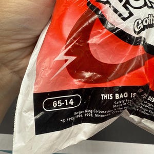 Op de afbeelding: Een gedeeltelijk zichtbare rode en witte plastic zak met de tekst "Pok" en "Gotta". De zak heeft een witte bliksemflits en de nummers "65-14" in een witte ovaal. De tekst "THIS BAG IS" is ook zichtbaar.