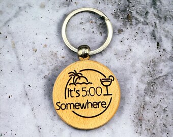 May include: Wooden keychain with a round, light brown tag. The tag has a black and white design of a palm tree, an island, and a cocktail glass. The text "It's 5:00 Somewhere" is engraved on the tag.