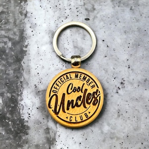 Może przedstawiać: Drewniany brelok do kluczy z okrągłą, brązową zawieszką z napisem "Official Member Cool Uncles Club". Brelok ma srebrny metalowy pierścień.