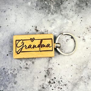 May include: Wooden keychain with a heart and the word "Grandma" engraved on it. The keychain is attached to a silver metal ring.