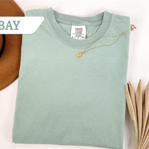 Pode incluir: Uma camiseta verde-claro dobrada com um colar dourado. A camisa tem uma etiqueta "Comfort Colors". Um chapéu marrom e folhas secas estão no fundo. A palavra "BAY" está em uma caixa branca no canto superior esquerdo.