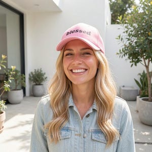 May include: A pink baseball cap with the word "blessed" embroidered in black. The cap is worn by a person wearing a light blue denim jacket. The background includes potted plants and a white wall.