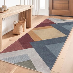 May include: A geometric patterned runner rug in shades of red, blue, gray, and tan. The rug is placed on a hardwood floor in a hallway, with a wooden bench, baskets, and a potted plant visible in the background. Two straw hats hang on the wall.