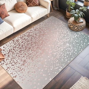 Puede incluir: Una alfombra rectangular con un diseño degradado, que transiciona de rosa claro a gris claro. La alfombra presenta un patrón de pequeñas formas dispersas. La alfombra está en un salón con un sofá blanco y cojines decorativos.