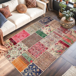 Op de afbeelding: Een woonkamer met een witte bank, kussens en een patchwork vloerkleed. Het vloerkleed bestaat uit vierkanten in rood, groen, blauw en beige. De kamer is ingericht met planten en een ingelijste wanddecoratie, wat een gezellige sfeer creëert.