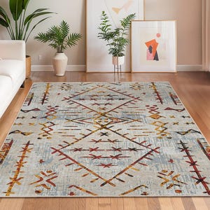 Puede incluir: Una alfombra rectangular con un patrón geométrico en un salón. La alfombra presenta un fondo gris claro con un diseño en rojo, naranja y marrón. La habitación incluye un sofá blanco, plantas y obras de arte enmarcadas.