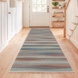 Könnte beinhalten: Ein langer, rechteckiger Teppich mit einem gestreiften Muster in Blau-, Rot- und Brauntönen. Der Teppich liegt in einem Flur mit Holzböden und weißen Wänden. Das Design des Teppichs verleiht dem Raum einen Hauch von Farbe und Textur.