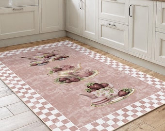 May include: A rectangular kitchen rug with a pink background and a white and pink checkered border. The rug features a repeating pattern of illustrations of teacups, strawberries, and pastries. The rug is placed on a wooden floor in a kitchen setting.
