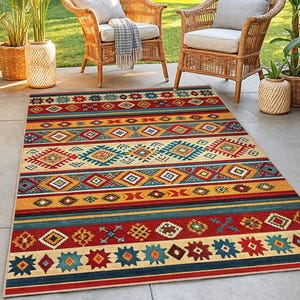 May include: A rectangular outdoor rug with a Southwestern design. The rug features a pattern of horizontal stripes in red, yellow, teal, and cream. Two wicker chairs with cushions are in the background, along with potted plants.