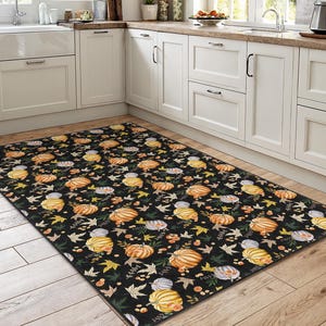 May include: A rectangular black rug with a repeating pattern of pumpkins, leaves, and berries in shades of orange, yellow, and green. The rug is placed in a kitchen setting with white cabinets and a wooden floor.