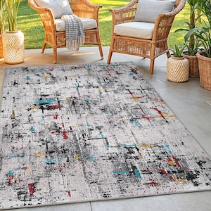 Puede incluir: Una alfombra rectangular para exteriores con un diseño abstracto. La alfombra presenta un fondo gris claro con un patrón de pinceladas negras, grises, rojas, amarillas y turquesas. Dos sillas de mimbre y plantas en macetas están en el fondo.