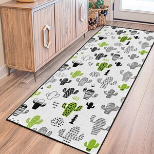 May include: A rectangular white rug with a black border featuring a repeating pattern of black and green cacti in various designs. The rug is placed on a wooden floor near a wooden cabinet and a white door.