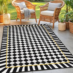 Puede incluir: Una alfombra de exterior a cuadros blancos y negros con un borde amarillo. La alfombra es rectangular y está colocada en un patio con dos sillas de mimbre y plantas en macetas en el fondo. El diseño de la alfombra presenta un patrón geométrico audaz.