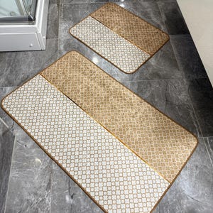 May include: Two bathroom rugs with a geometric pattern. The larger rug is beige and white with a geometric pattern. The smaller rug is beige with a geometric pattern.