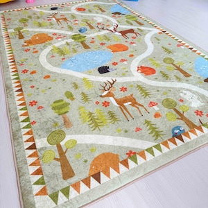 Könnte beinhalten: Eine grüne und braune Spielmatte mit einem gewundenen Pfad und verschiedenen Waldtieren, darunter Hirsche, Bären und Vögel. Die Matte hat einen beigefarbenen Rand mit einem dreieckigen Muster.