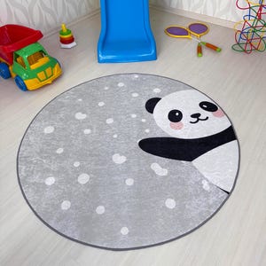 Op de afbeelding: Een rond grijs tapijt met een wit pandabeerontwerp. De pandabeer glimlacht en heeft zwarte ogen en een zwarte neus. Het tapijt is bezaaid met witte stippen.