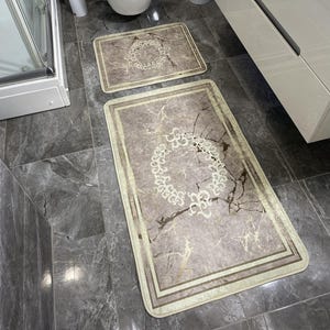 May include: Two beige bathroom rugs with a marble pattern and a gold-colored design. The larger rug is placed in front of a white toilet and the smaller rug is placed in front of a white sink.