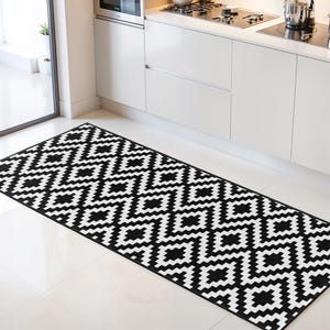 Puede incluir: Una alfombra rectangular con un patrón geométrico en blanco y negro. La alfombra está colocada sobre un suelo de baldosas blancas frente a un mueble de cocina blanco. El diseño de la alfombra presenta un patrón de diamantes repetido.
