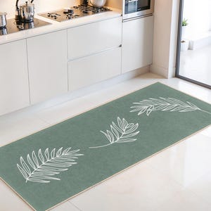 Puede incluir: Una alfombra de cocina rectangular verde salvia con un borde crema. La alfombra presenta tres diseños de hojas blancas estilizadas. La alfombra está colocada frente a un mueble de cocina blanco y una estufa de acero inoxidable.