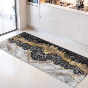 Könnte beinhalten: Ein rechteckiger Küchenteppich mit Marmormuster. Der Teppich hat eine Mischung aus Schwarz, Grau und Gold. Die goldenen Akzente erzeugen einen schimmernden Effekt und verleihen dem Bodenbelag einen Hauch von Eleganz. Der Teppich liegt vor einer weißen Küchenzeile.