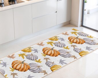 Puede incluir: Alfombra de cocina rectangular con un patrón repetido de calabazas naranjas, amarillas y grises, junto con hojas de otoño, sobre un fondo blanco. La alfombra está colocada frente a un mueble de cocina blanco.