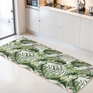 Può includere: Un tappeto da cucina rettangolare con un design ad acquerello di grandi foglie tropicali verdi. Le foglie hanno diverse tonalità di verde e bianco, creando un'estetica lussureggiante e naturale. Il tappeto è posizionato su un pavimento piastrellato bianco.