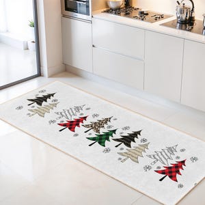Op de afbeelding: Een rechthoekig kersttapijt met een herhalend patroon van gestileerde kerstbomen in verschillende kleuren en patronen, waaronder ruitjes en luipaardprint, met sneeuwvlokken op een witte achtergrond. Het tapijt is geplaatst in een keuken.