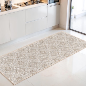 Puede incluir: Una alfombra de cocina rectangular con un estampado floral beige y crema. La alfombra está colocada frente a un mueble de cocina blanco y una estufa. El diseño de la alfombra presenta un patrón repetido de flores estilizadas y otras formas.