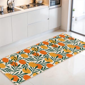 May include: A rectangular kitchen rug featuring a repeating pattern of orange and yellow fruit and green leaves on a cream background. The rug is positioned in front of white kitchen cabinets.