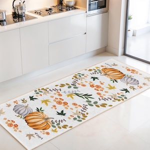 May include: A rectangular kitchen rug featuring a fall-themed design. The rug has a cream-colored background with a border of pumpkins, leaves, and flowers in shades of orange, green, and brown. The rug is placed in front of a white kitchen cabinet.