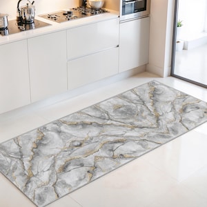 Peut inclure: Un tapis de cuisine rectangulaire avec un motif de marbre. Le tapis présente une palette de couleurs grise et blanche avec des veines dorées. Le tapis est placé devant un meuble de cuisine et une cuisinière blancs.